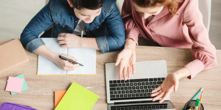 Top view of homeschool mother and son sitting at desk with laptop and notebooks and studying homeschool curriculum together