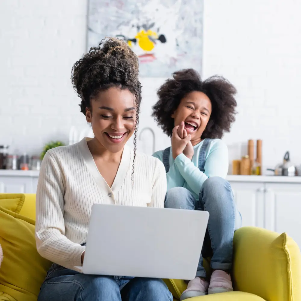 mom writing review on laptop smiling on couch with elementary aged child laughing